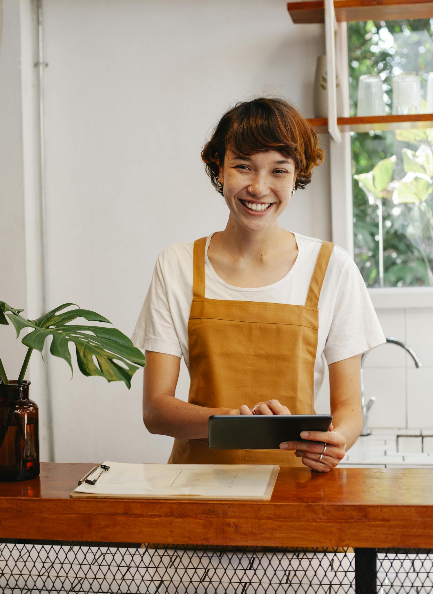 Cheerful barista using a tablet in a charming café, next to a plant and clipboard.
