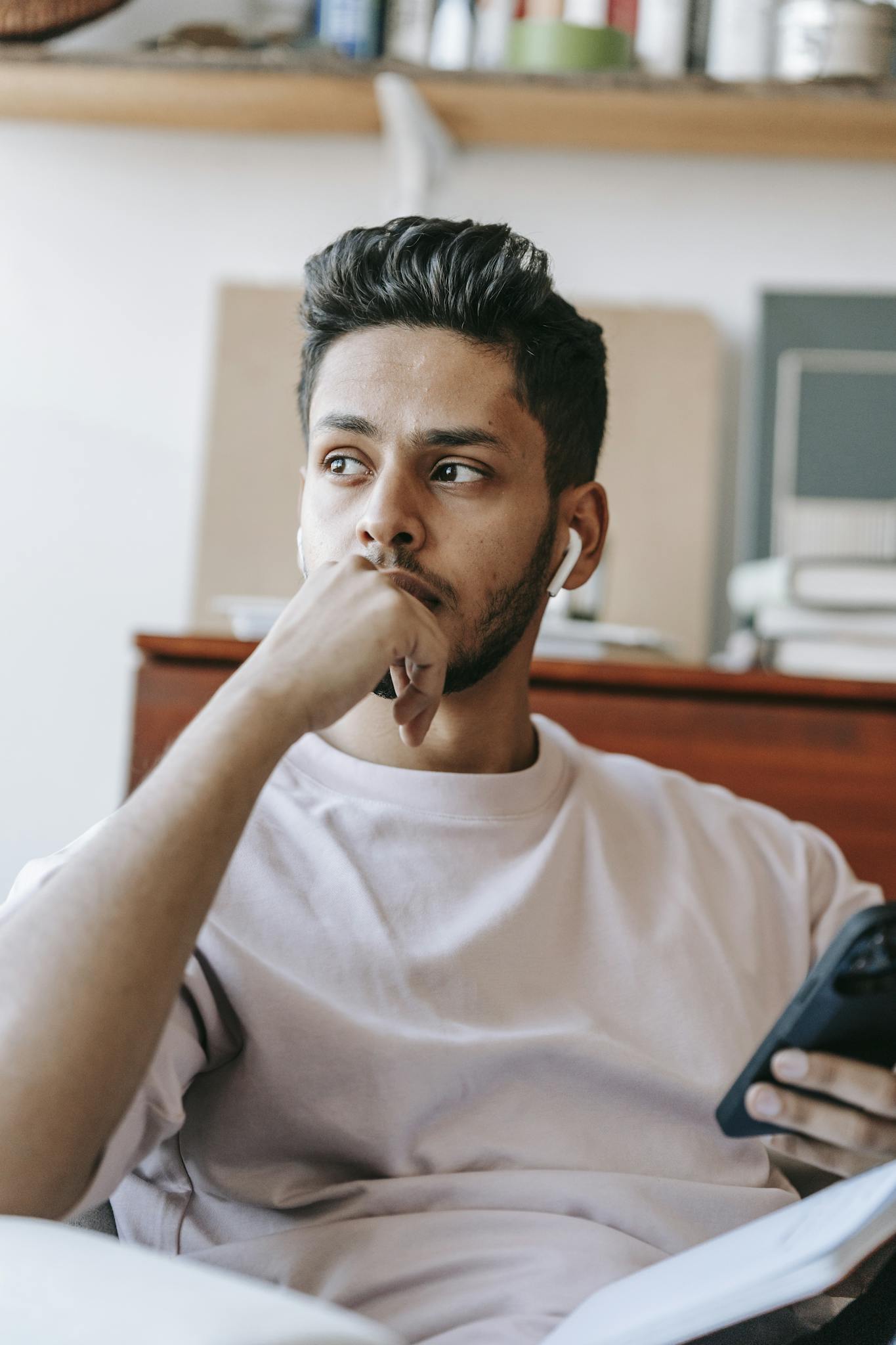 Pensive young ethnic male wearing casual outfit and earbuds browsing cellphone and looking away thoughtfully while sitting on armchair and touching chin